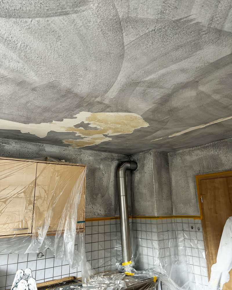 Eine stark verrußte Küche mit deutlichen Rauch- und Brandschäden an Decke und Wänden. Die Decke zeigt großflächige graue Rußspuren und abgeplatzte Stellen, an den Schränken und Fliesen hängen Schutzfolien. Ein Abluftrohr ragt aus der Wand, und auf der Arbeitsfläche liegen Reste von Putz und Ruß. Die Szene dokumentiert den Zustand nach einem Küchenbrand und den bevorstehenden Sanierungsbedarf.