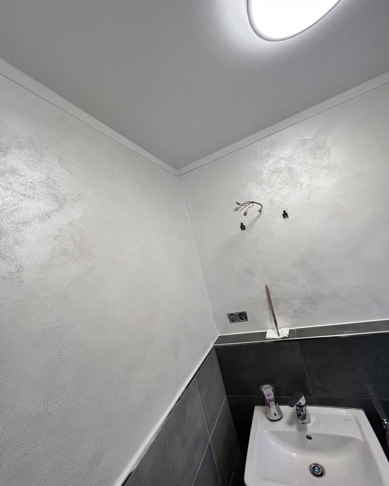 Ein frisch renoviertes Bad mit hell verputzten Wänden und sauber gearbeiteten Übergängen zur Decke. Der obere Wandbereich wurde neu beschichtet, während der untere Bereich aus dunklen Fliesen besteht. Über dem Waschbecken sind Installationskabel für die spätere Montage einer Lampe sichtbar. Die Szene zeigt präzise Putz- und Malerarbeiten im Sanitärbereich.