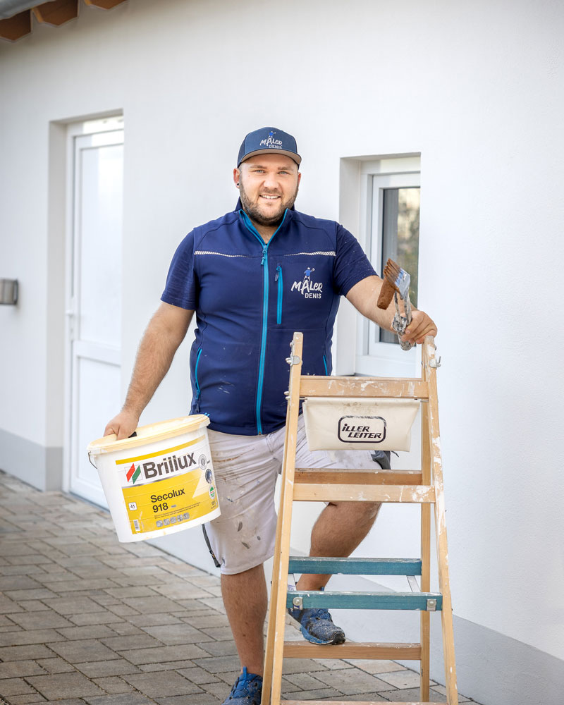 Ein Maler steht vor einer frisch renovierten Fassade und hält einen Farbeimer sowie einen Pinsel in der Hand. Er trägt Arbeitskleidung und eine Kappe mit Firmenlogo. Neben ihm befindet sich eine mit Farbresten versehene Stehleiter. Die Szene zeigt typische Vorbereitungen für Außenanstriche und vermittelt ein professionelles, handwerkliches Arbeitsumfeld.